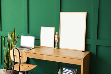 Modern workplace with laptop, mannequin and blank frames near green wall