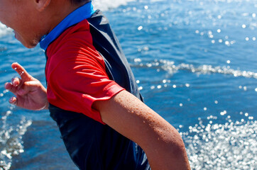 Happy smiling boy running on the beach. Enjoying nice summer vacations. Blue and bright sea. 
Wearing of a Rashguard swim shirt, swimwear. Protection of the skin from UV rays.