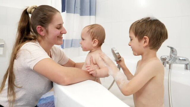 Older boy helping his mother washing baby brother in bath with shower head. Concept of children hygiene, healthcare and family care at home