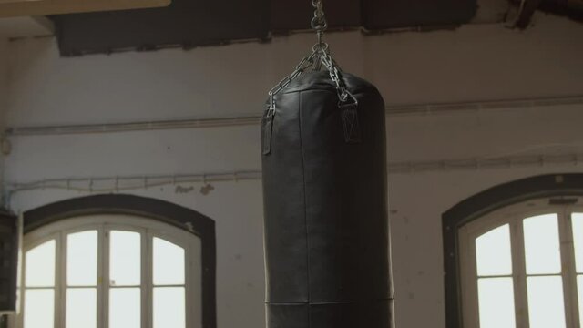 Medium shot of leather heavy bag hanging from ceiling in gym. Horizontal shot of punching bag swinging in empty boxing gym in daytime. Sport, workout, training concept