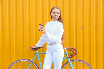 Pretty teenage girl with bicycle and cup of coffee near yellow fence