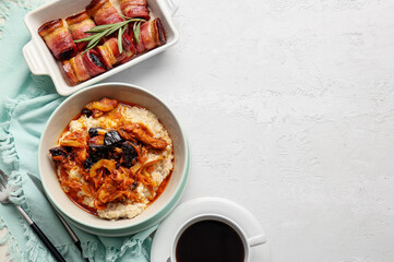 Bowl with delicious oatmeal, beef stew and  prunes baked in crispy bacon and rosemary branch in baking dish on light background