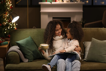 Little African-American girl and her mother with cup of hot cocoa reading book on Christmas eve