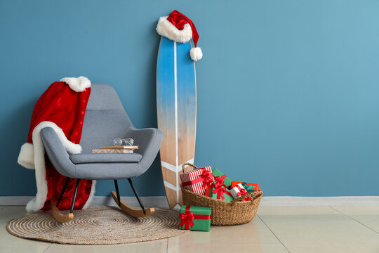 Armchair with Santa Claus suit, blue surfboard and basket with Christmas gifts near color wall