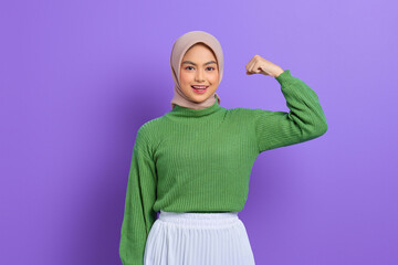 Beautiful smiling Asian woman in green sweater raises arms and shows biceps, demonstrated her achievement after training in a gym isolated over purple background