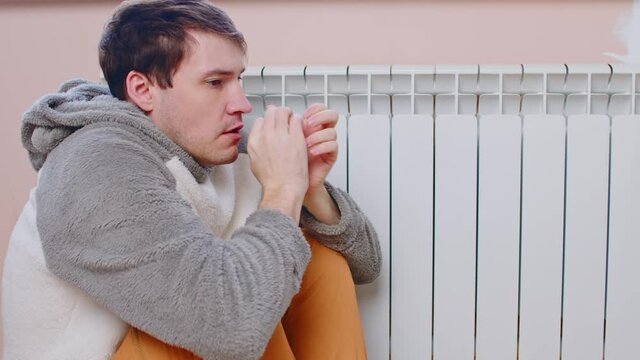 Man sitting on floor and warming near radiator. Frozen male cringing from cold, warming near heating radiator in winter day.