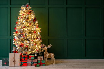 Beautiful Christmas tree with glowing lights, gifts and wooden reindeer near green wall