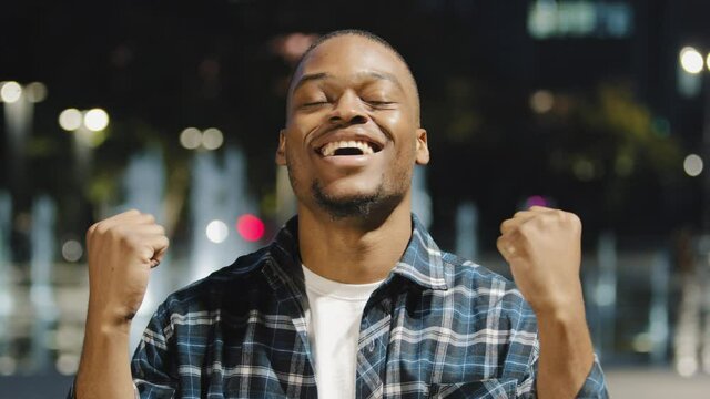 Male portrait african american man guy in plaid shirt stands in city in evening at night looking at camera says wow rejoicing win victory good luck winning gesture with hands talking, triumph concept