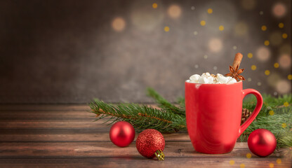 Red mug of warm drink with marshmallows and green Christmas tree branches with red Christmas ornaments on dark wooden background