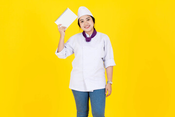 Portrait studio shot of Asian professional female executive chef in white cooking uniform with hat and scarf standing smiling holding learning new recipes from notebook in hands on yellow background