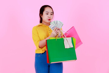 Fototapeta premium Portrait studio shot of Asian young rich wealthy female shopaholic shopper in casual outfit smiling holding hundred dollars fan shopping bags on pink background