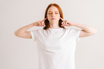 Portrait of calm young woman covering her ears and gesticulating say no bla-bla-bla standing on white isolated background in studio. Irritated redhead female showing nonsense content.