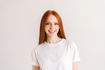 Portrait of positive pretty young woman smiling and looking at camera, standing on white isolated background in studio. Happy cheerful redhead female showing sincere positive emotion.