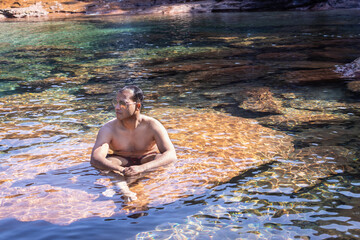 young man wearing sun glass sitting on rock in flowing river clear water at morning from flat angle