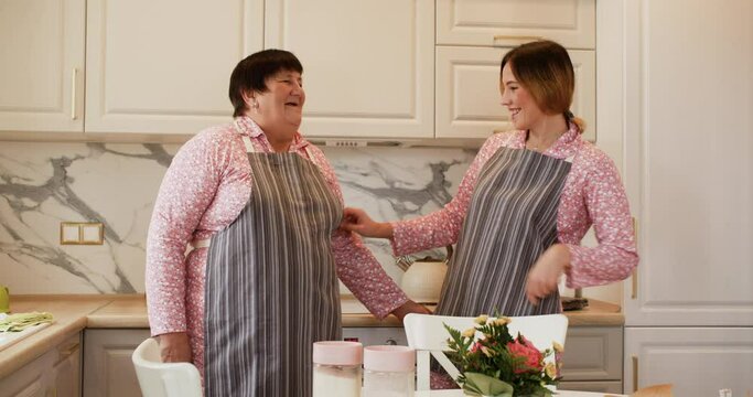 Happy housewives wearing matching clothes and cooking aprons on home kitchen. Smiling adult daughter and senior mother in culinary aprons on cozy kitchen