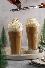 Grating chocolate over two tall glasses with cold coffee drink frappe - iced cappuccino with whipped cream, christmas decoration and trees