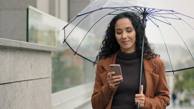 Portrait of beautiful happy cheerful young brunette girl with long black curly hair woman with phone and transparent umbrella walking in city street near building looking at smartphone chatting online