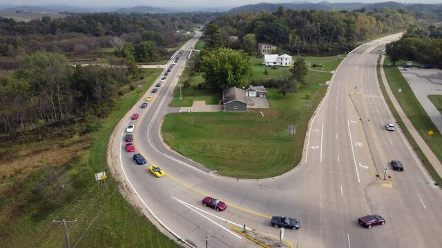 Corvette car parade through rural roads in Wisconsin. Cars turning onto highway. Residential home and garage on corner. Multiple directional signs present. 