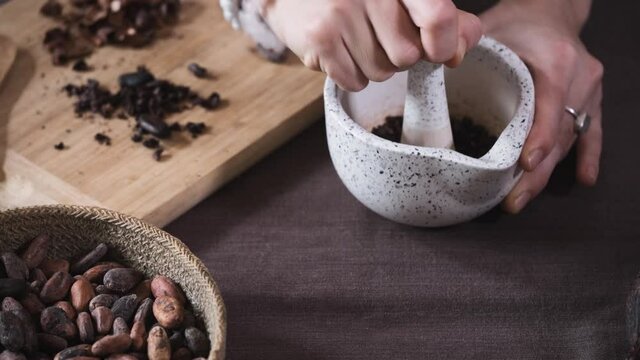 Woman hands putting organic cacao beans in pounder on table, cocoa nibs, artisanal chocolate making in rustic boho style for ceremony. Degustation, Chocolate making with pounder close-up top view