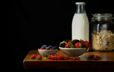 Still life of berry fruits on wooden table with bottles of milk and cereals against black background
