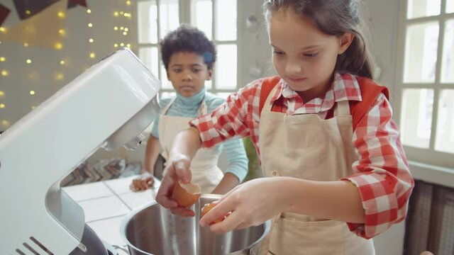 Little Asian and Caucasian girls cracking eggs and adding them to mixer bowl while cooking with biracial boy during culinary masterclass