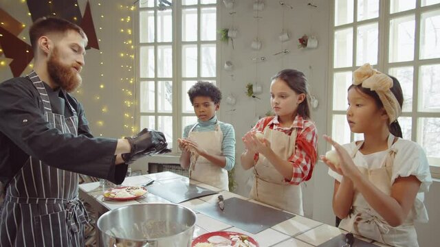 Group of cute multiethnic kids looking at chef and rolling dough into shape while learning baking on cooking masterclass