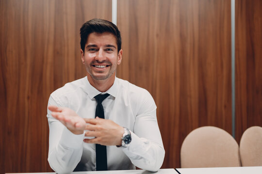 Young Businessman With Laptop At Table In Office Looking At Camera And Pointing Hands Gesture. Video Conference Call Web Cam Meeting.