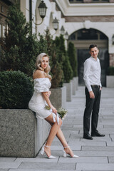 Fototapeta premium A beautiful blonde bride with curly hair with a bouquet of peonies in her hands sits near the green thuja, cypress against the background of the groom standing in the city. Wedding photography.