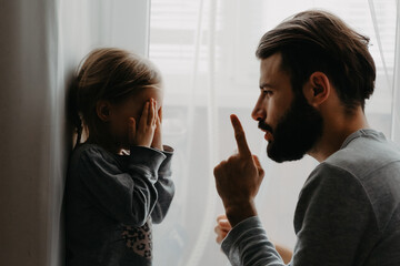 The father puts the child in a corner and makes remarks for bad behavior by waving his index finger near his face. The child does not want punishment and apologizes - obrazy, fototapety, plakaty
