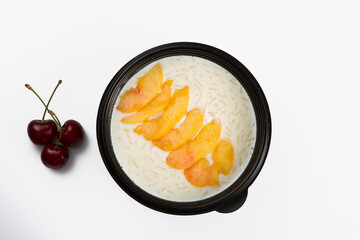 isolated close up top view shot of a delicious white rice milk porridge with peach fruit slices on top in a black plastic bowl and cherries on a white background