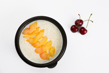 isolated close up top view shot of a delicious white rice milk porridge with peach fruit slices on top in a black plastic bowl and cherries on a white background