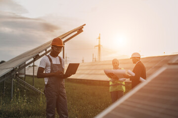 African american technician in uniform standing on field with solar panels and typing on laptop...