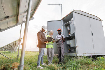 Indian man and muslim woman with clipboard and laptop talking with african american technician at solar station. Multiracial people examining process of green energy production.