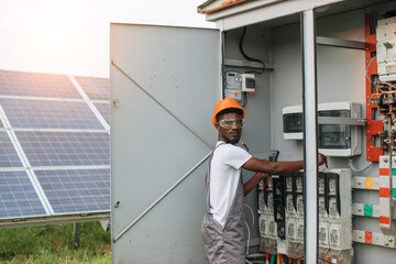 African american man in grey overalls and orange helmet repairing cables in switchgear at solar station. Concept of people, maintenance and alternative energy.