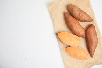 Parchment with cut and whole sweet potatoes on white table, top view. Space for text