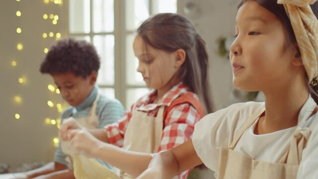 Little Asian girl preparing dough for baking, smiling and telling something during kids cooking class