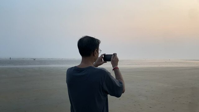 Cinematic parallax shot of a man holding phone and clicking picture of the beautiful sunset in a empty sea beach.