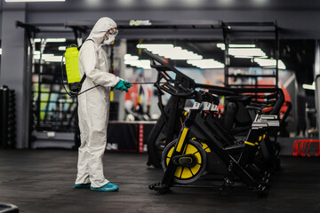 Fototapeta premium Coronavirus outbreak situation. A male person in a special protective suit refreshes and cleans fitness equipment in a spinning cycling gym spot. Keep distance in public to protect from COVID-19