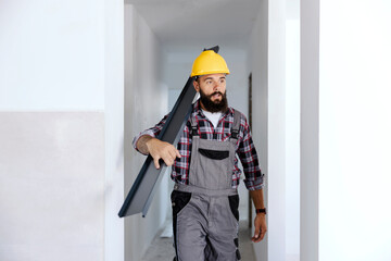 A strong handyman with a helmet on his head is relocating materials in a new apartment. He is holding a metal beam.