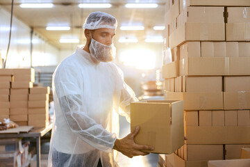 A food factory worker in a white sterile uniform is holding an opened box with cookies and...