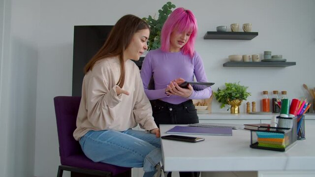 Lovely pink haired young woman and charming female student with congenital disability and leg prosthesis discussing university report, doing research online using digital tablet in college dorm room.