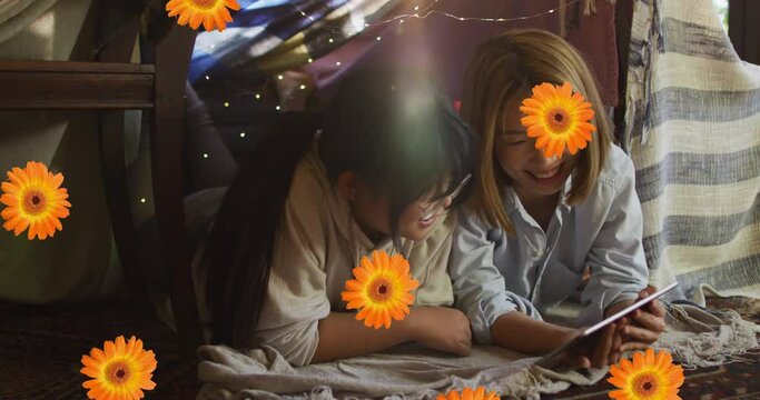 Animation of orange flowers over happy asian mother and daughter using tablet at home