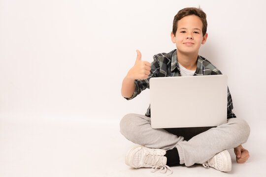 Smiling Cute Boy Wearing Plaid Shirt Sitting On Floor Holding Laptop Computer With Thumb Up Isolated Over White Background.