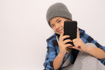 Cute boy wearing casual clothing sitting on the floor looking to camera while using smartphone isolated over white background.
