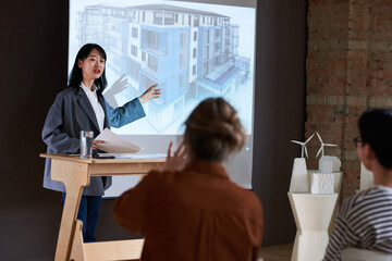 Young businesswoman pointing at architectural blueprint and presenting new project to people during a seminar