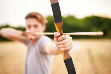Young male sportsman targeting with traditional bow - Teenager archer practicing archery in nature - Outdoors sports and recreation concept with a millennial boy - Focus on the archer's hand - obrazy, fototapety, plakaty