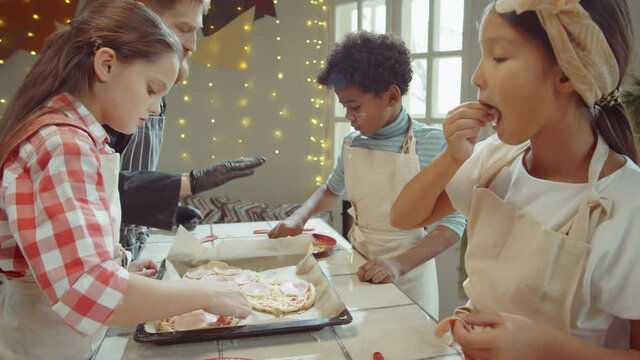 Group of multiethnic children adding ham to pizza before baking with assistance of male chef during culinary class