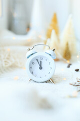 The alarm clock behind the Christmas garlands of bokeh in the house on the table with a sweater in the background and decorations. Winter mood, festive decoration, magical Christmas.