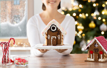 winter holidays, baking and food cooking concept - close up of happy smiling woman holding gingerbread house at home over christmas tree lights background