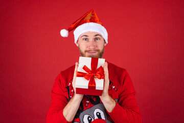 Red-bearded man in a santa hat and a christmas sweater on a red background with a gift box is excited, looking forward to the holiday